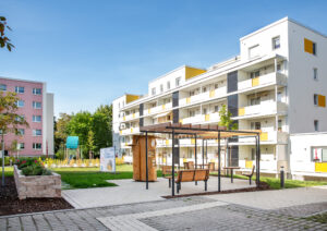 Bücherbaum im Kappler Stadtgarten, umgeben von einer überdachten Sitzecke, bepflanzten Hochbeeten. Im Hintergrund ist der Spielplatz und die Wohnhäuser zu sehen.